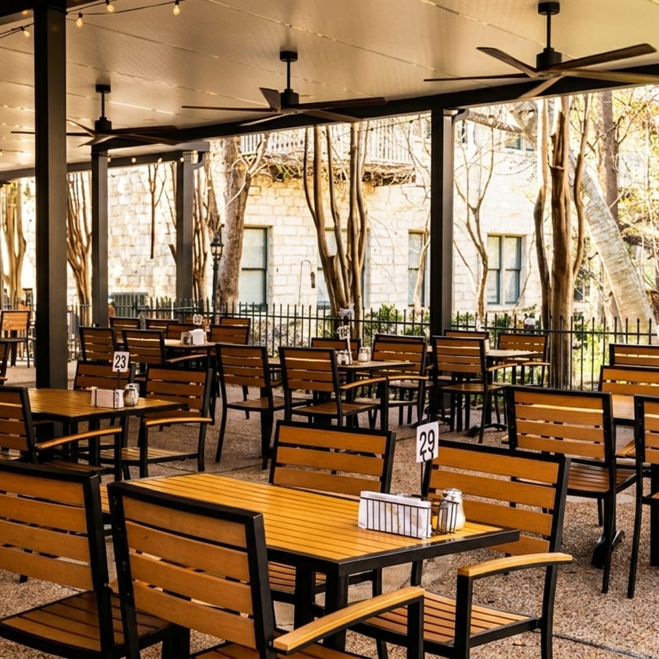 An empty outdoor patio with wooden tables and chairs, string lights, and ceiling fans.